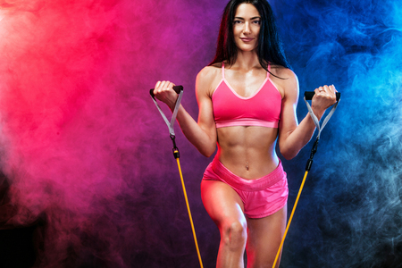 Muscular young fit sports woman athlete. Workout with bands or expander in gym on black background with colo smoke. Studio shot.の写真素材