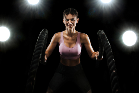 Battle ropes. Fit sports woman, athlete working out in functional training doing exercise on black background. Fitness, gym and workout motivation.の写真素材