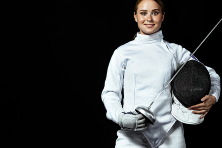 Young fencer athlete wearing fencing costume holding the sword and mask. Isolated on black backgroundの写真素材