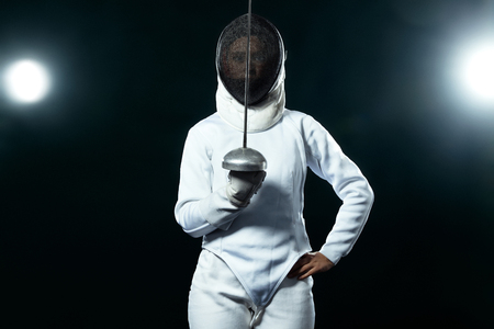 Young fencer athlete wearing fencing costume holding the sword and mask. Isolated on black background with lightsの写真素材