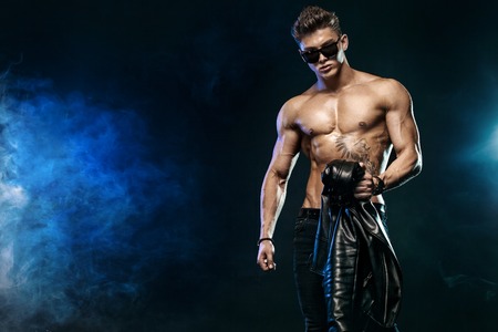 Men fashion. Close-up portrait of a brutal and fit man topless in sunglasses and leather jacket....