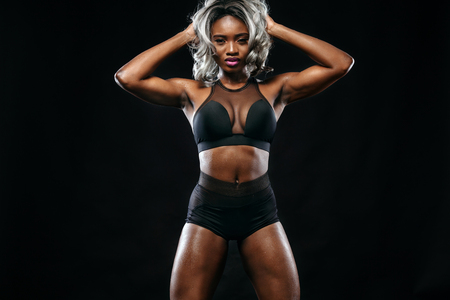 Sporty beautiful afro-american bodybuilder model, woman in sportwear makes fitness exercising at black background to stay fitの写真素材