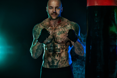 Sportsman with tattoos, man boxer fighting in gloves with boxing punching bag. Isolated on black background with smoke. Copy Space.の写真素材
