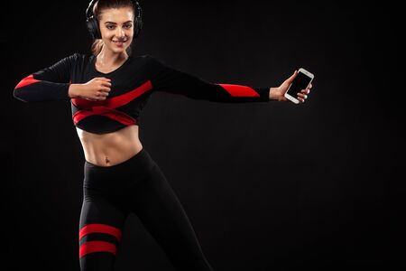 Beautiful young and fit woman in sportswear and headphones listening music from phone and smiling. Isolated on a black background.の写真素材