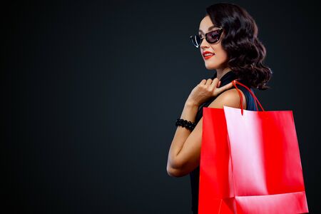 Black Friday sale concept for shop. Shopping woman in sunglasses holding red bag isolated on dark background.の写真素材
