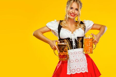 Wow emotion. Young sexy Oktoberfest girl waitress, wearing a traditional Bavarian or german dirndl, serving big beer mugs with drink isolated on yellow background.の写真素材