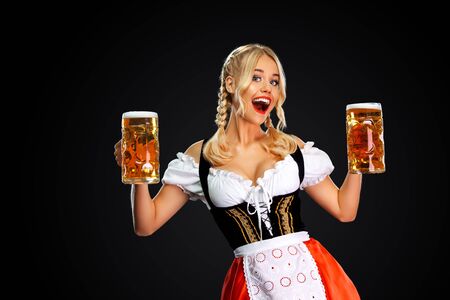 Happy sexy oktoberfest girl waitress, wearing a traditional Bavarian or german dirndl, serving two big beer mugs with drink isolated on black background.の写真素材