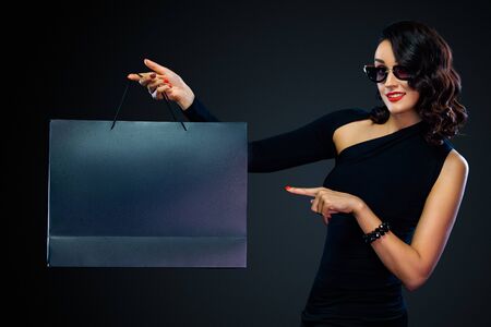 Black Friday sale concept for shop. Girl in sunglasses holding big bag isolated on dark background at shopping. Woman pointing to looking on copy space for sales text.の写真素材