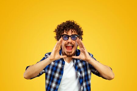 Closeup portrait of handsome happy european man with beard and curly hair in blue shirt opened mouth widely and shouting loud. Hispter isolated on yellow background. Positive emotions.の写真素材
