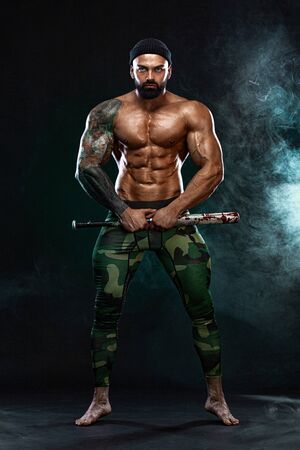 Strong and fit man bodybuilder with baseball bat shows abdominal muscles under a t-shirt. Sporty muscular guy athlete. Sport and fitness concept. Mens fashion.の写真素材