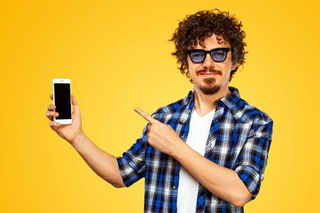 European man with curly hair in blue sunglasses with mobile phone or smarphone. Guy pointing to looking on the screen. Handsome stylish hipster in plaid shirt posing over yellow background.の写真素材
