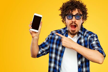 European man with curly hair in blue sunglasses pointing to looking on the screen of mobile device or smarphone. Handsome stylish hipster in plaid shirt posing over yellow background.の写真素材