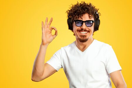 Portrait of happy bearded man with curly hair pointing ok sign with fingers isolated over yellow background. Guy in blue sunglasses pick on great idea.の写真素材