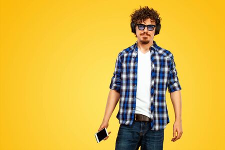 European man with curly hair in blue sunglasses with mobile phone or smarphone. Handsome smiled stylish hipster in plaid shirt posing over yellow background.の写真素材