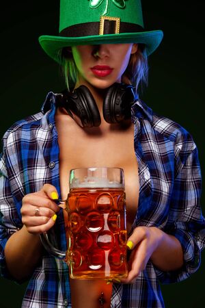 Saint Patrick Day. Young sexy Oktoberfest girl waitress in green har serving big beer mugs with beer isolated on dark background. St Patricks day celebration.の写真素材