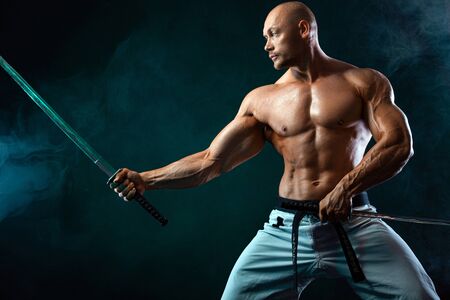 Karate fighter on black background with smoke. Shirtless man samurai with Japanese sword. Fit man...
