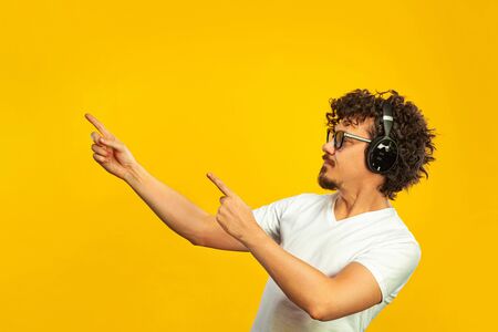 Portrait of happy bearded man with curly hair pointing finger at screen isolated over yellow background. Guy in blue sunglasses and headphones pick on great idea.の写真素材