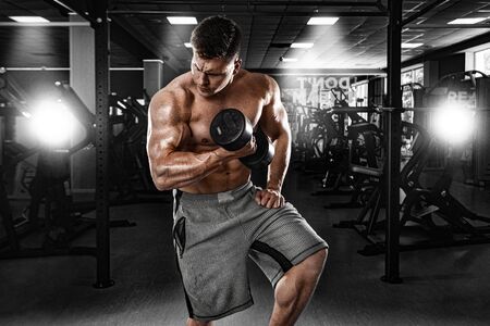 Brutal strong muscular bodybuilder athletic man pumping up muscles with dumbbell in the gym. Workout bodybuilding concept. Copy space for sport nutrition ads.の写真素材