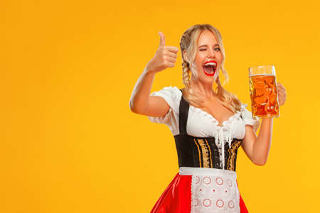 Young sexy Oktoberfest girl waitress, wearing a traditional Bavarian or german dirndl. Woman serving big beer mugs with drink isolated on yellow background.の写真素材