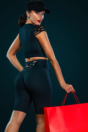 Black friday sale concept for fitness shop. Shopping woman in sportswear and hat holding red bag isolated on dark backgroundの写真素材