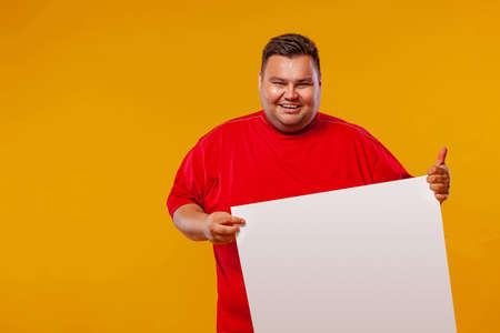Fat man in red t-shirt displaying a white banner ad isolated over a yellow background.の写真素材