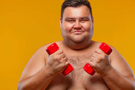 Fat man doing workout traning with dumbbells. Happy guy isolated over yellow background.の写真素材