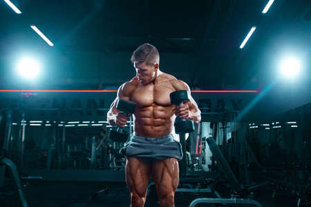Bodybuilder athlete man pumping up muscles in the gym with dumbbells. Brutal strong muscular guy on fitness workout. Bodybuilding concept.の写真素材
