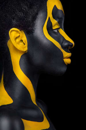 Face art. Woman with black and yellow body paint. Young african girl with colorful bodypaint. An amazing model with yellow makeup. Closeup face.