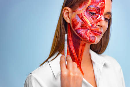 Cosmetic injection in the face. Young woman with half of face with muscles structure under skin. Model for medical training on a light background.の写真素材