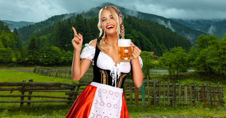 Oktoberfest girl waitress with beer. Woman wearing a traditional Bavarian or german dirndl on octoberfest, serving big mugs with drink on mountains background. Woman pointing to looking up.の写真素材