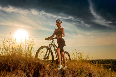 Biker-girl at the sunset with mountain bike. Woman with bicycle at sunrise on trail.の写真素材