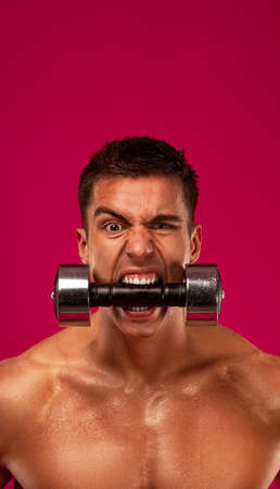 Bodybuilding concept. Brutal strong muscular bodybuilder athletic man pumping up muscles with dumbbell on red background. Workout bodybuilding concept. Copy space for sport nutrition ads.の写真素材