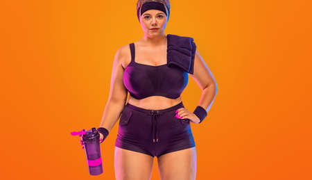 The importance of rest after gym. Girl recovering from cardio. Size plus model woman with with towel and protein shaker in sportswear on orange background. Sports and healthy lifestyleの写真素材