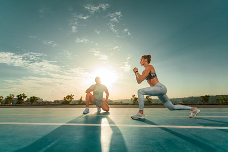 Instructor and athlete runner on the track. The athletics marks the time in the lunges exercise on a stopwatch. Fitness trainer and mentee.の写真素材