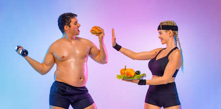 Fat man chooses between burger and fitness with dumbbells. Trainer girl tells him stop and gives vegetables instead of fast food. Not sporty men eating hamburger isolated on pink background.の写真素材