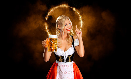 Smiling young sexy  girl waitress, wearing a traditional Bavarian or german dirndl, serving big beer mug with drink isolated on black background.の写真素材