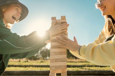 Tower game. Happy family with kid playing board games outdoors in the park. Smiling mom and teen girl sitting on the floor and pulling wooden blocks from the towerの写真素材