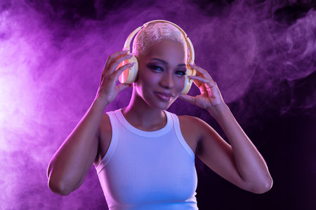 Hipster fashion girl, african american model wear headphones enjoy listen new cool music mix. Woman stand at purple studio background in trendy club purple party light with fog.の写真素材