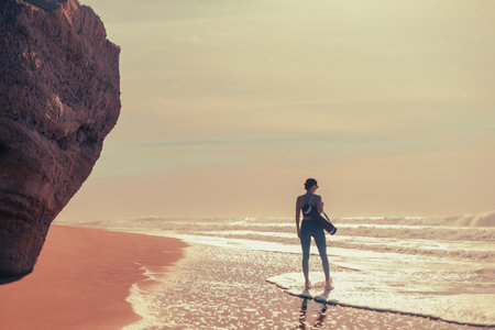 Fit woman walking by the ocean with yoga mat perfect for wellness and coastal lifestyle themes. Ideal for yoga retreats or fitness promotions.の写真素材
