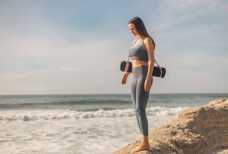 Peaceful moment before or after yoga session as woman relaxes by the water holding her mat on a sunny coast.の写真素材