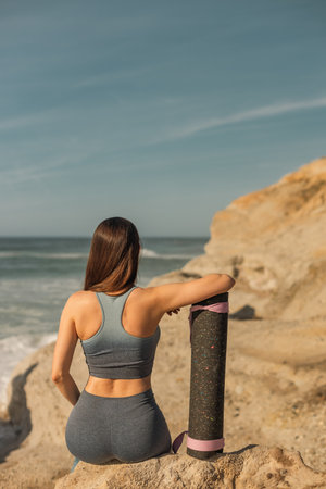 Slim girl sitting by the seaside holding a yoga mat great for promoting active outdoor living and yoga brandsの写真素材