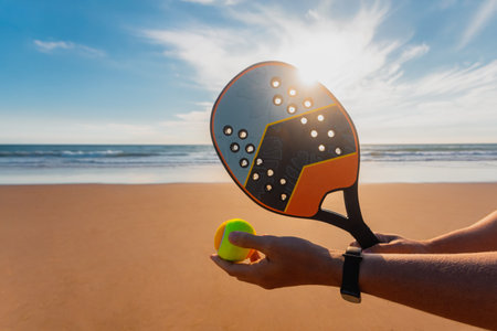 Beach tennis. Closeup photo of hands with racket and ball. Professional beach tennis player hitting the ball on sandy shore during sunset training.の写真素材