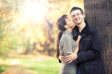 Pretty young girl kisses in cheek her boyfriend in the park; autumn leaves and sunbeams on the...