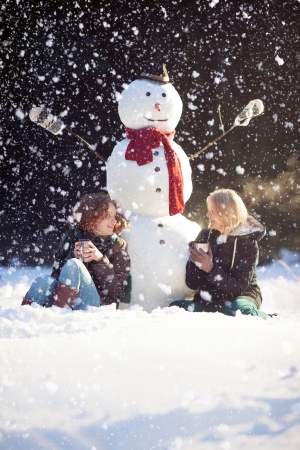 Two young woman sitting near snowman and drinking hot tea while itの写真素材