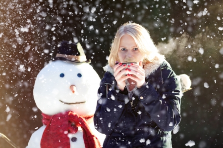 Pretty blond girl dinking tea outdoors with snowman while it's snowingの写真素材