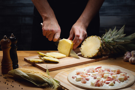 Chef cooking hawaiian pizza, cutting fresh pineapple. Low key shot, close up of hands, some...