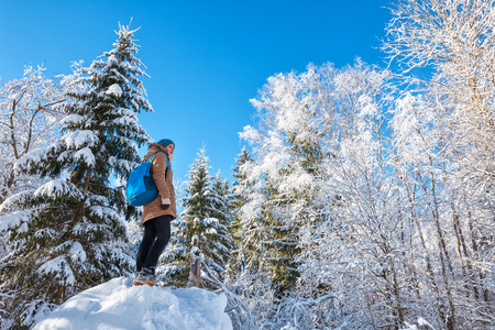 Young woman with blue backpack hiking in winter forest, enjoying the view from the top of snowdrift