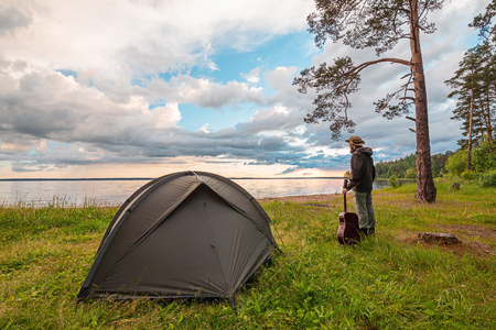 Tourist with guitar standing near camp tent on lake shore, enjoying the view at sunset. Unity with nature.の写真素材