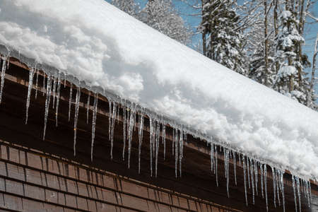Wooden cabin with snowy roof and icicles at sunny winter day, close up