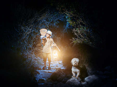 A young naturalist caught something into a jar. At dark the boy walks out of a cave with a lantern and a butterfly net, examines his catch. His faithful dog is nearby.の写真素材
